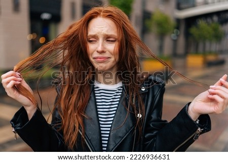 Portrait of sad attractive redhead young woman touching wet hair after autumn rain standing on beautiful city street. Front view of unhappy lady untangling hair after being caught in rain. Royalty-Free Stock Photo #2226936631