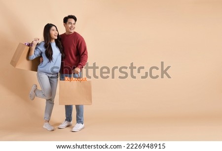 Happy smiling Asian couple with shopping bags in sweater enjoying their winter discount sale isolated on beige color background. Shopaholic fashionable 20s man and woman looking away with copy space.