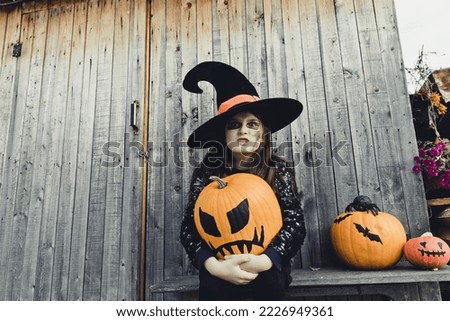 Little girl in Halloween costume of witch sitting on bench with orange pumpkin jack-o-lanterns on wooden wall copy space  background