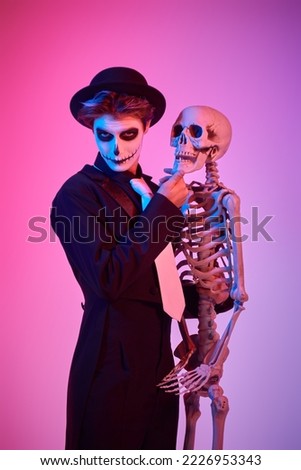 Portrait of a teenage boy in a costume of skeleton posing with a human skeleton at studio in mixed colored lighting. Halloween party for teenagers. 