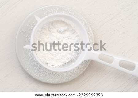 Icing sugar in a plastic sieve on top of white ceramic plate on white wood. Top view.