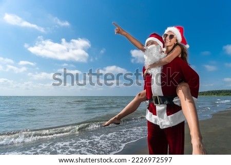 Happy Santa Claus is holding on his back a smiling young woman in a Santa hat with a pointing finger to the side on the seashore on a sunny day. Celebrating Christmas holidays in warm countries