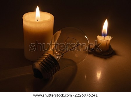 Switched off light or not glowing light bulb liying near a burning candles in total darkness. Blackout city, electricity off, load shedding, energy crisis or power outage, symbolic image. Royalty-Free Stock Photo #2226976241