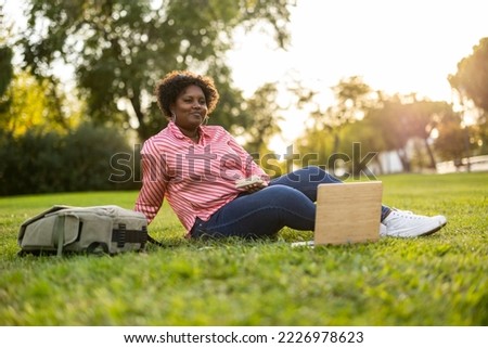 Young african american curvy female eats a sandwich and studying in a park with the laptop sitting on the grass at sunset