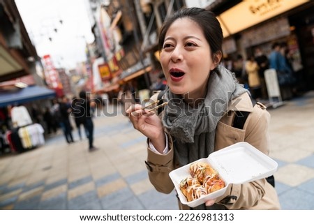 dutch angle shot of cheerful Asian Japanese girl making funny face expression while eating hot delicious takoyaki octopus balls at a food plaza in Shinsekai Osaka Japan