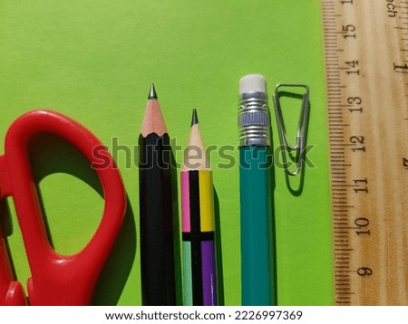 Red rings of scissors, three simple pencils, a paper clip and a wooden ruler in the sun, on a light green background (macro, top view, slightly tilted diagonally, fragment).