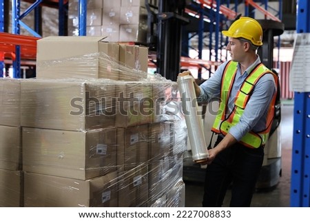 Caucasian warehouse worker man with hardhat and reflective jackets wrapping boxes in stretch film parcel on pallet while control stock and inventory in retail warehouse logistics distribution center