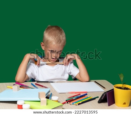 Boy attentively looks at phone with his mouth open, holds brush with his hands, paints, an album, pencils, books, cubes on table. Selective focus. Learning to draw, hobbies in your free time.
