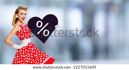Woman holding paper heart with % sign.  Portrait image of happy pin up girl. Blond model at retro fashion rockabilly sales ad. Valentine or like symbol. Black Friday sale. Blurred mall background. 