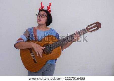 An Asian woman wearing a Christmas-themed headband plays a guitar and sings carols; happy expression.