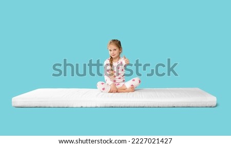 Little girl sitting on mattress and showing thumb up against light blue background