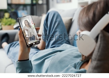 Woman lying down on the couch at home and watching social media videos on her smartphone Royalty-Free Stock Photo #2227026727