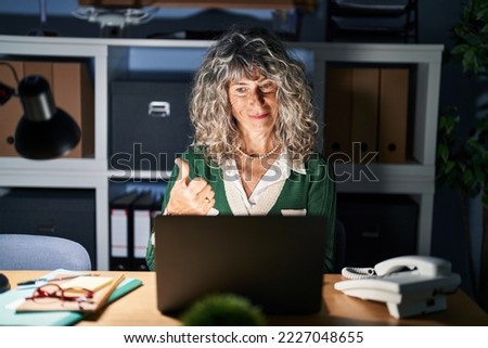 Middle age woman working at night using computer laptop doing happy thumbs up gesture with hand. approving expression looking at the camera showing success. 