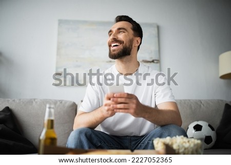 cheerful man using smartphone near bottle of beer and tasty food on blurred foreground Royalty-Free Stock Photo #2227052365
