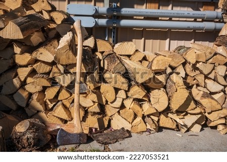 A still-life of a stack of chopped wood and an axe along the side of a wall with the sun shining upon it. 