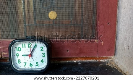 Stock photo of square shape retro style black color alarm clock kept near window, window covered by mosquito net. Picture captured under natural light at Gulbarga, Karnataka, India. focus on object.