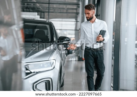 Standing and holding black graphic tablet in hand. Young man in white clothes is in the car dealership.