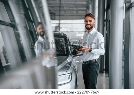 Standing and holding black graphic tablet in hand. Young man in white clothes is in the car dealership.