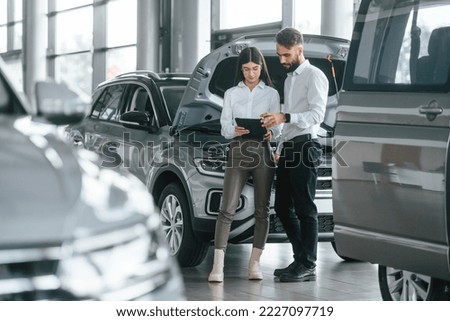 Reading information by using tablet. Man with woman in white clothes are in the car dealership together. Royalty-Free Stock Photo #2227097719