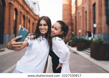 Positive young female friends taking self shot on smartphone while standing in street of old town smiling happily and looking at screen