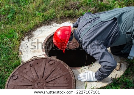 A male plumber opened the hatch of a water well and looks inside. Inspection of water pipes and meters. Royalty-Free Stock Photo #2227107101
