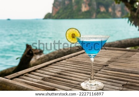 A glass of blue hawaii cocktail alcohol drink decorated with lime with the sea in background.