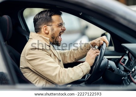 An annoyed businessman in smart casual is honking on other drivers and yelling in a car. Royalty-Free Stock Photo #2227122593