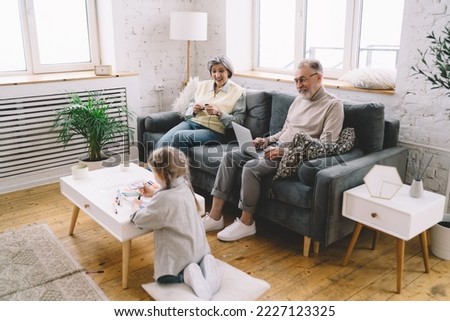 Elderly man using netbook and woman knitting while sitting on couch spending family time with little girl drawing at table