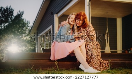 Beautiful Teenage Granddaughter Hugging with Her Grandmother While Using a Smartphone. Girl Showing Family Photos and Videos to Her Grandparent. Relatives Sitting Outside on a Porch on a Warm Day. Royalty-Free Stock Photo #2227127777