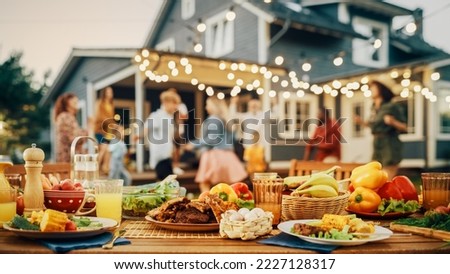 Outdoors Dinner Table with Gorgeous-Looking Barbecue Meat, Fresh Vegetables and Salads. Happy Joyful People Dancing to Music, Celebrating and Having Fun in the Background on Home Porch. Royalty-Free Stock Photo #2227128317