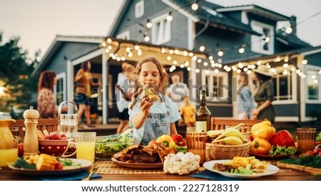 Outdoors Dinner Table with Delicious Barbecue Meat and Fresh Vegetables and Salads. Little Girl Eating a Grilled Corn. Happy People Dancing and Having Fun in the Background. Royalty-Free Stock Photo #2227128319