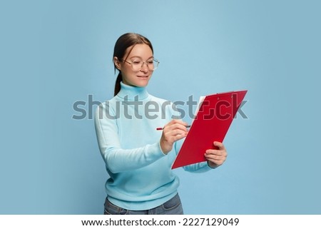 One young woman writing down on paper, passing test, exam, or signing documents, contract isolated over blue background. Concept of youth culture, emotions, facial expression, fashion, occupation