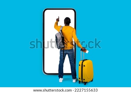 Back view of african american man traveller standing next to big smatphone with white empty touch screen, carrying yellow luggage, holding passport with tickets, mockup, blue studio background Royalty-Free Stock Photo #2227155633