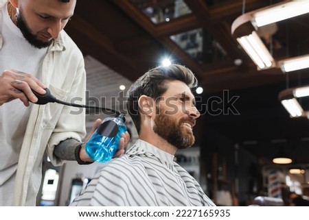 bearded man smiling near barber spraying perfume on his hair Royalty-Free Stock Photo #2227165935