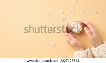 Winter season skin care cosmetics concept. First person top view photo of woman's hands in knitted sweater small cream jar and snowflakes on isolated pastel beige background with empty space