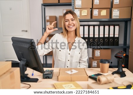 Young blonde woman working at small business ecommerce smiling and confident gesturing with hand doing small size sign with fingers looking and the camera. measure concept. 