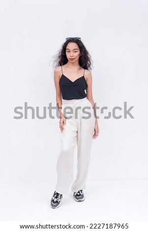 A slim and pretty Filipino woman with long curly hair in her early 20s. Wearing a black spaghetti strap blouse and white pants. Isolated on a white background.