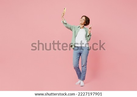 Full body young woman she wear green shirt white t-shirt doing selfie shot on mobile cell phone post photo on social network show v-sign isolated on plain pastel light pink background studio portrait
