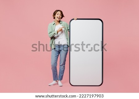 Full body young woman wear green shirt white t-shirt point finger on big huge blank screen mobile cell phone smartphone with workspace copy space mockup isolated on plain pastel light pink background