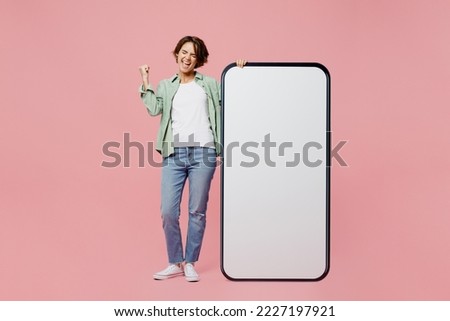 Full body young woman wear green shirt white t-shirt near big huge blank screen mobile cell phone smartphone with workspace mockup area do winner gesture isolated on plain pastel light pink background