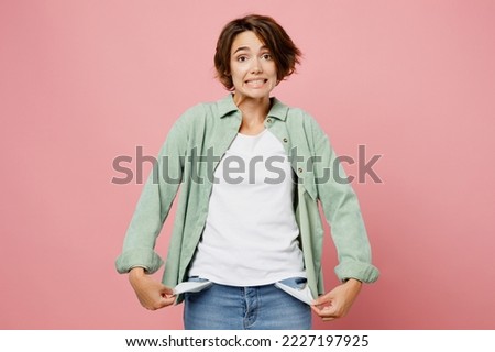 Young sad woman 20s she wear green shirt white t-shirt show empty pockets like she is pennieless and no money isolated on plain pastel light pink background studio portrait. People lifestyle concept
