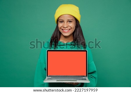 Little fun IT kid teen girl of African American ethnicity 13-14 years old wear casual hoody hat work on use laptop pc computer with blank screen workspace area isolated on plain dark green background