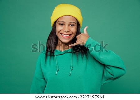 Little smiling happy kid teen girl of African American ethnicity 13-14 years old wear casual hoody hat do phone gesture like says call me back isolated on plain dark green background Childhood concept