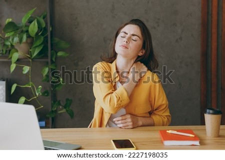 Young sad tired employee business woman 20s she wear casual yellow shirt put hand on shoulder neck close eyes suffer fro pain sit work at wooden office desk with pc laptop. Achievement career concept Royalty-Free Stock Photo #2227198035