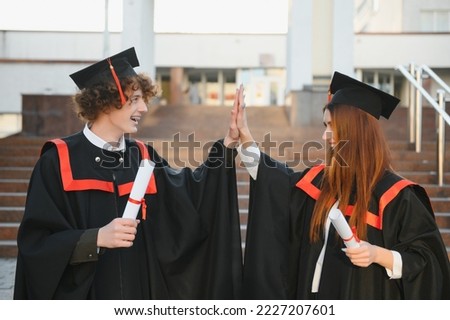 Two joyful students in black academic gowns and caps standing in a library among the bookshelves. Educational concept. Graduation. Modern generation