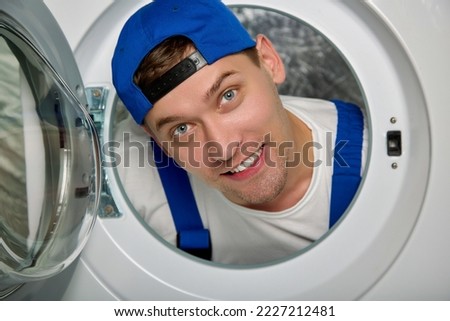 Close-up of the head of a man repairman of household appliances, poked through an open hole for loading laundry. Cheerful plumber repairs a washing machine