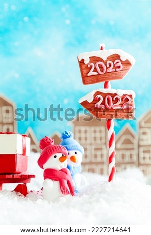 Two cute plasticine snowmen with sleds and gifts in a toy town. Wooden direction signs 2023 and 2022. Waiting for the New Year 2023, Christmas card.
