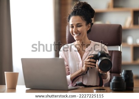 Happy creative millennail woman photographer working at office studio, sitting at workdesk with modern laptop on, holding professional camera, drinking coffee, checking photos, copy space