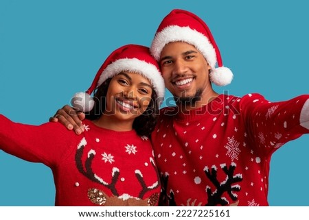 Happy beautiful black couple embracing, taking selfie while celebrating Christmas together, wearing Santa hats and red sweaters with deer, isolated on blue studio background, closeup photo