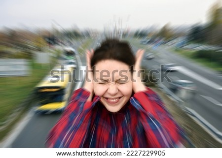 Woman with closed eyes covering her ears in the street while vehicles are passing by fast in the background. Stressful and frustrating situation. Noise pollution concept. Radial blur  filter applied. Royalty-Free Stock Photo #2227229905
